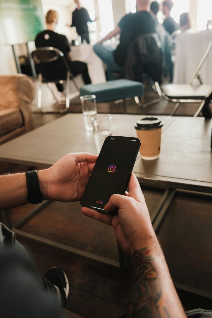 Tattooed hands hold a smartphone displaying the Instagram logo in a cozy indoor setting.